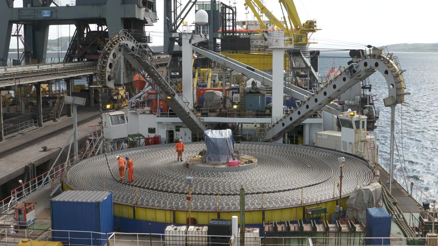 An industrial cable‑laying ship features a massive circular turntable filled with coiled subsea cable, with several workers in protective clothing standing on the platform and large mechanical arms positioned above it.