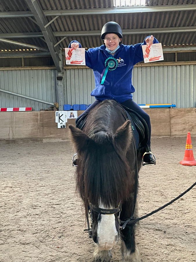 Young person holding up riding achievements while riding a horse.